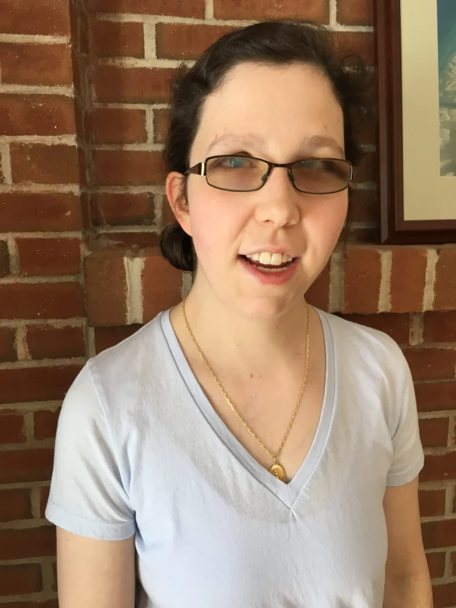 Picture of Jameyanne smiling in a v-neck shirt in front of a red brick wall.