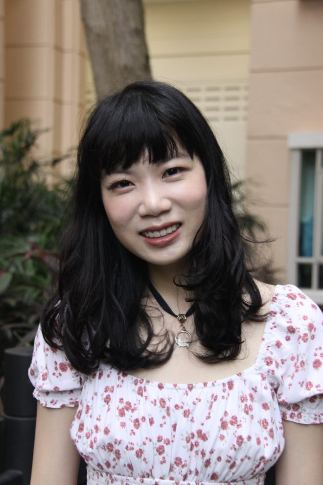 Megan Chee, smiling, her head tilted slightly to the right. She has long wavy dark hair and bangs, and is wearing a white dress with a red floral print.