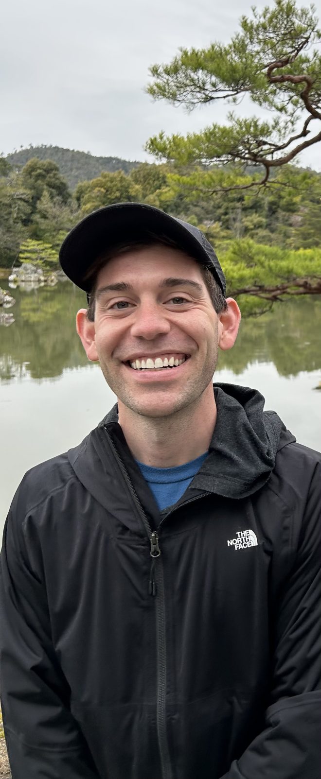 Author Ryan Cole, dressed in a dark baseball hat and raincoat, standing in front of a lake
