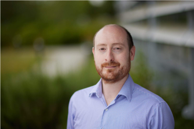 Head and upper torso of Isaac Harwood, who is a bearded man with balding red hair. He wears a pale blue shirt. He is standing in front of a blurred background of a building, lake and trees.