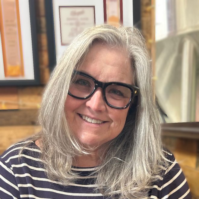 Stephannie Tallent is smiling at the camera in front of a bookshelf. She wears a black and white striped top and black-framed glasses. She has mid-length platinum hair