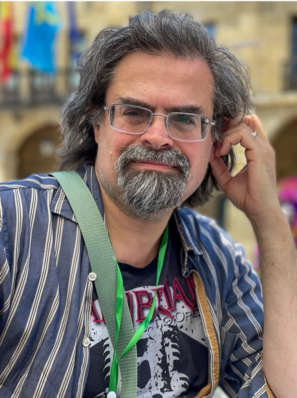 Adrian Tchaikovsky has salt-and-pepper shoulder length hair and a goatee beard. He wears a blue striped shirt, green braces, and clear-framed glasses. Photo by @catsparkx